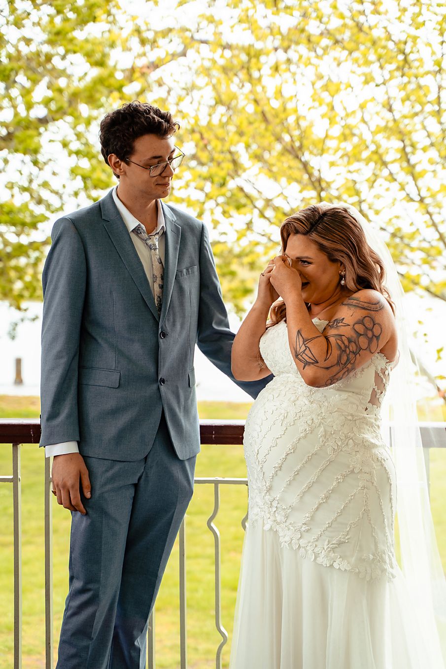 A couple in wedding attire share a playful moment on a balcony with autumn trees in the background.