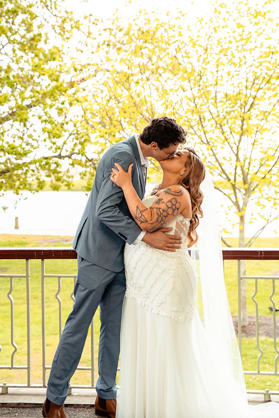 A romantic wedding couple shares an intimate kiss on an outdoor patio surrounded by autumn trees with golden leaves.