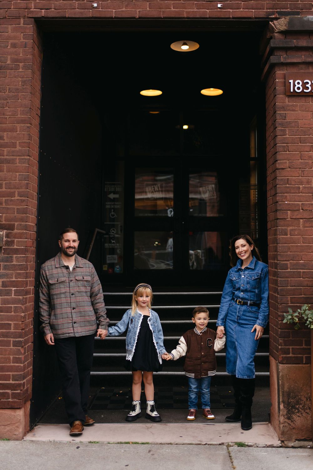 A family group stands together in front of a brick building entrance with recessed lighting.