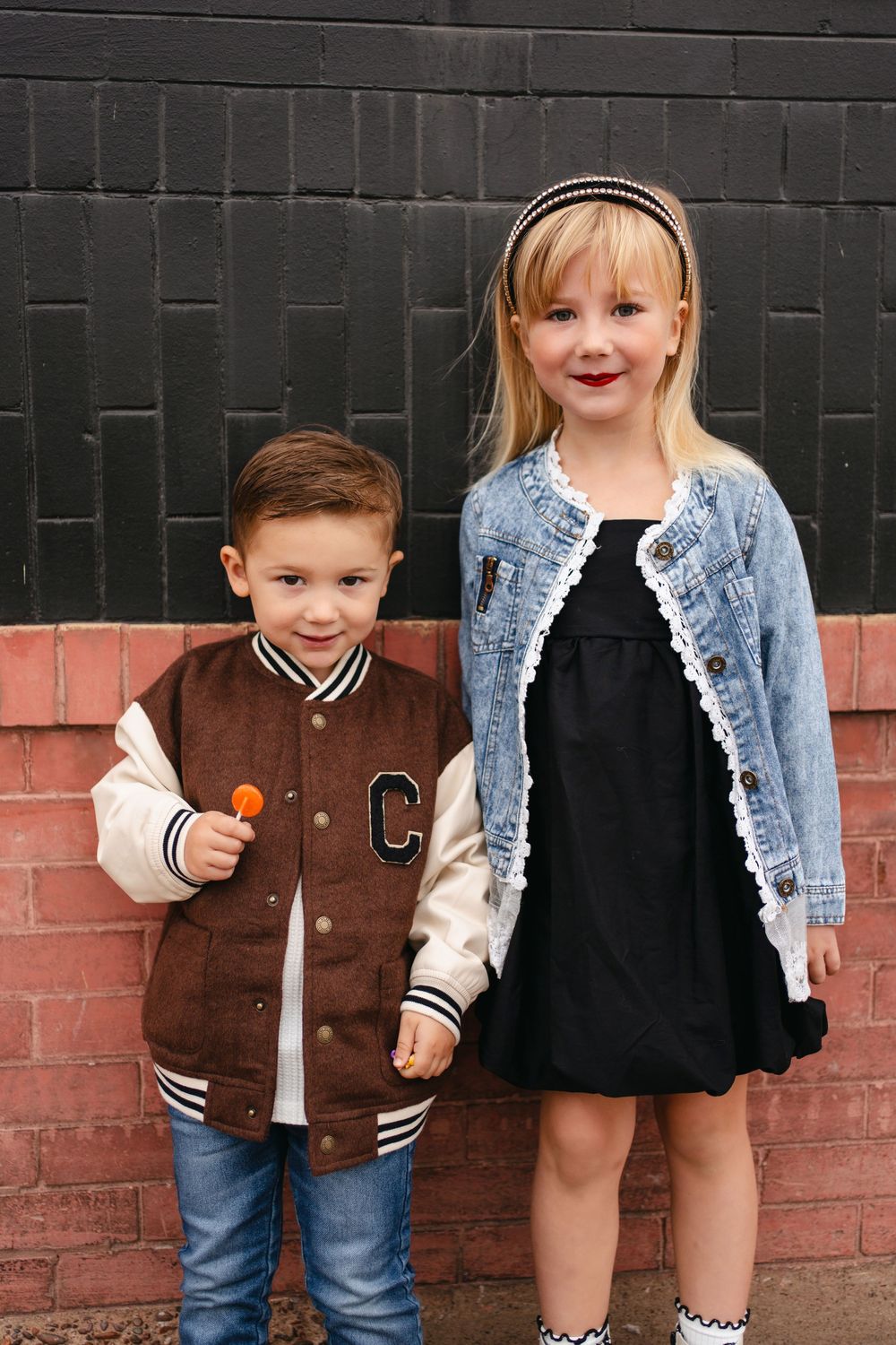 Two children stand together wearing casual outfits by a brick and black painted wall.