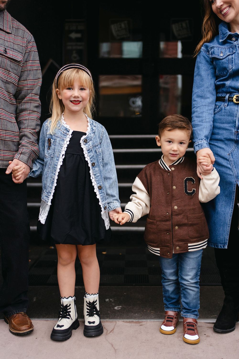 Two children hold hands with parents while wearing coordinated denim and varsity jacket outfits.
