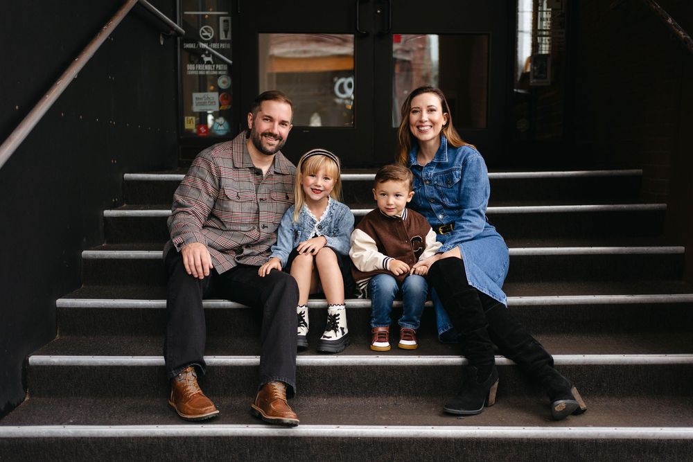 A family of four sits together on dark wooden steps, smiling warmly at the camera.