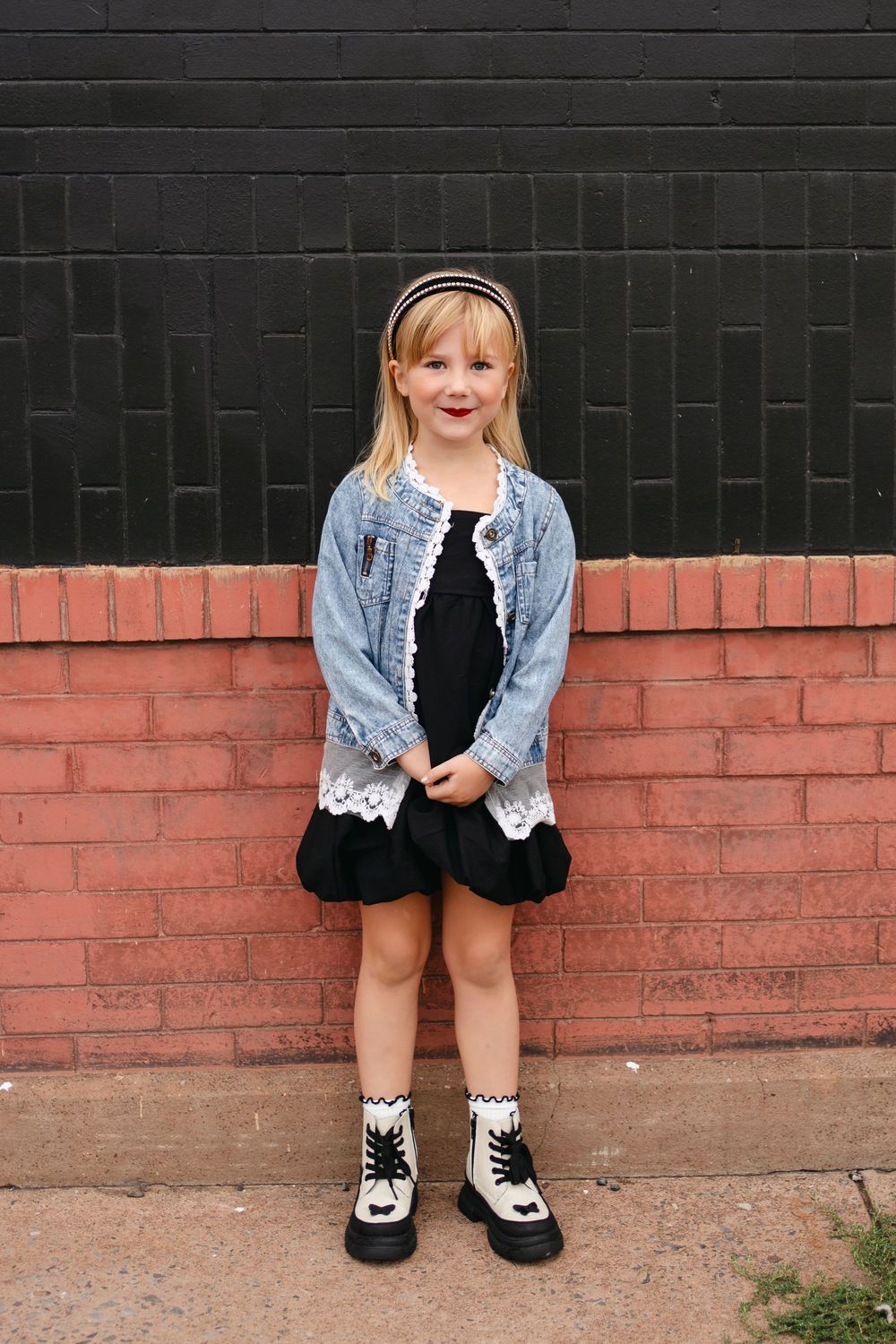 Child wearing denim jacket and black dress poses against contrasting black and red brick walls.