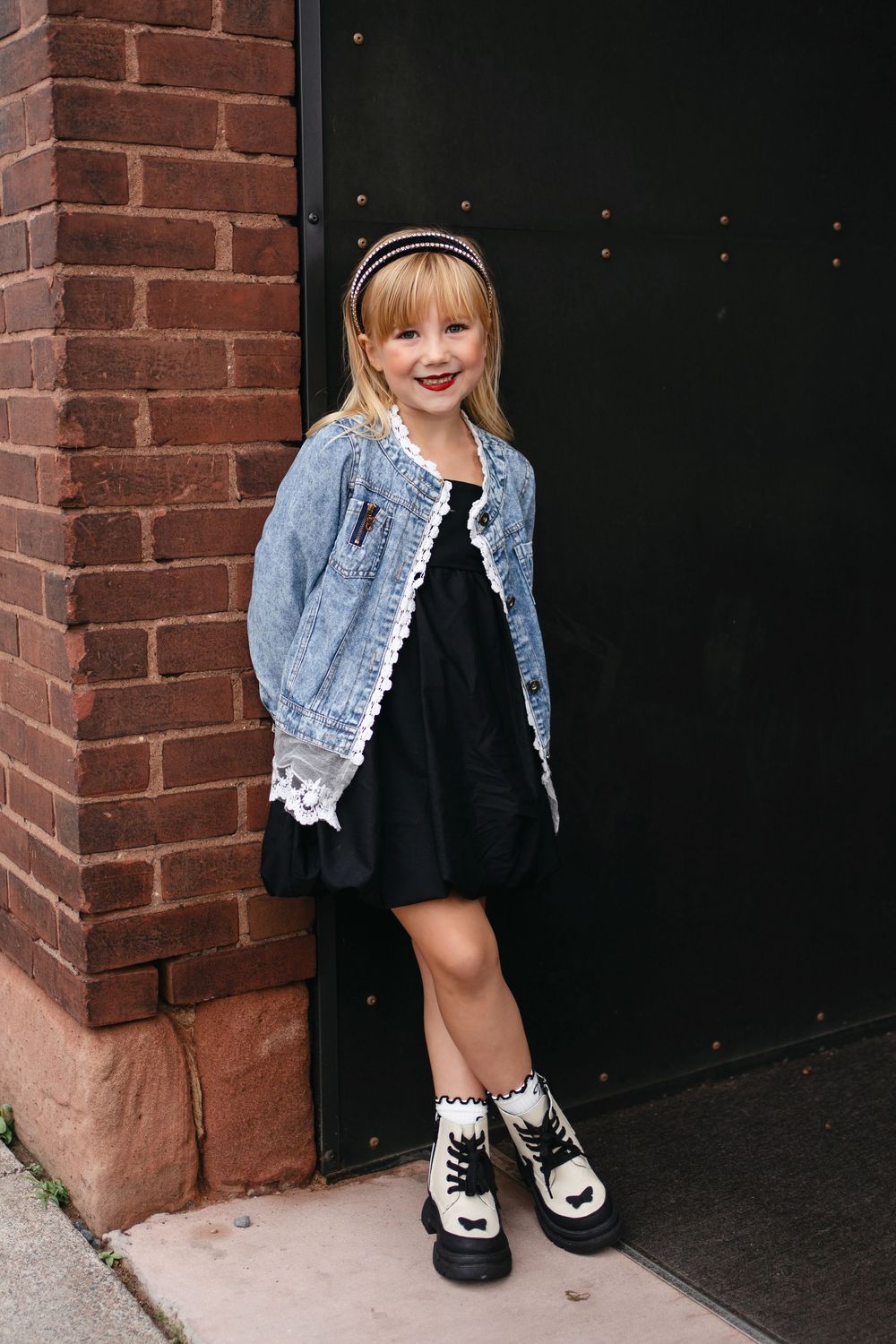 A child in a denim jacket and black dress poses near a dark doorway with brick walls.