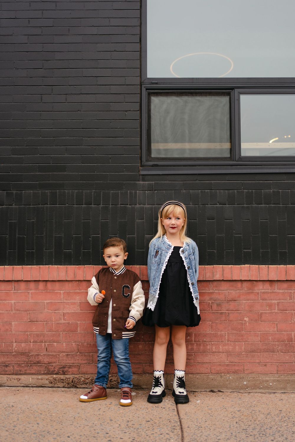 Two children stand against a black and red brick wall wearing stylish casual outfits.