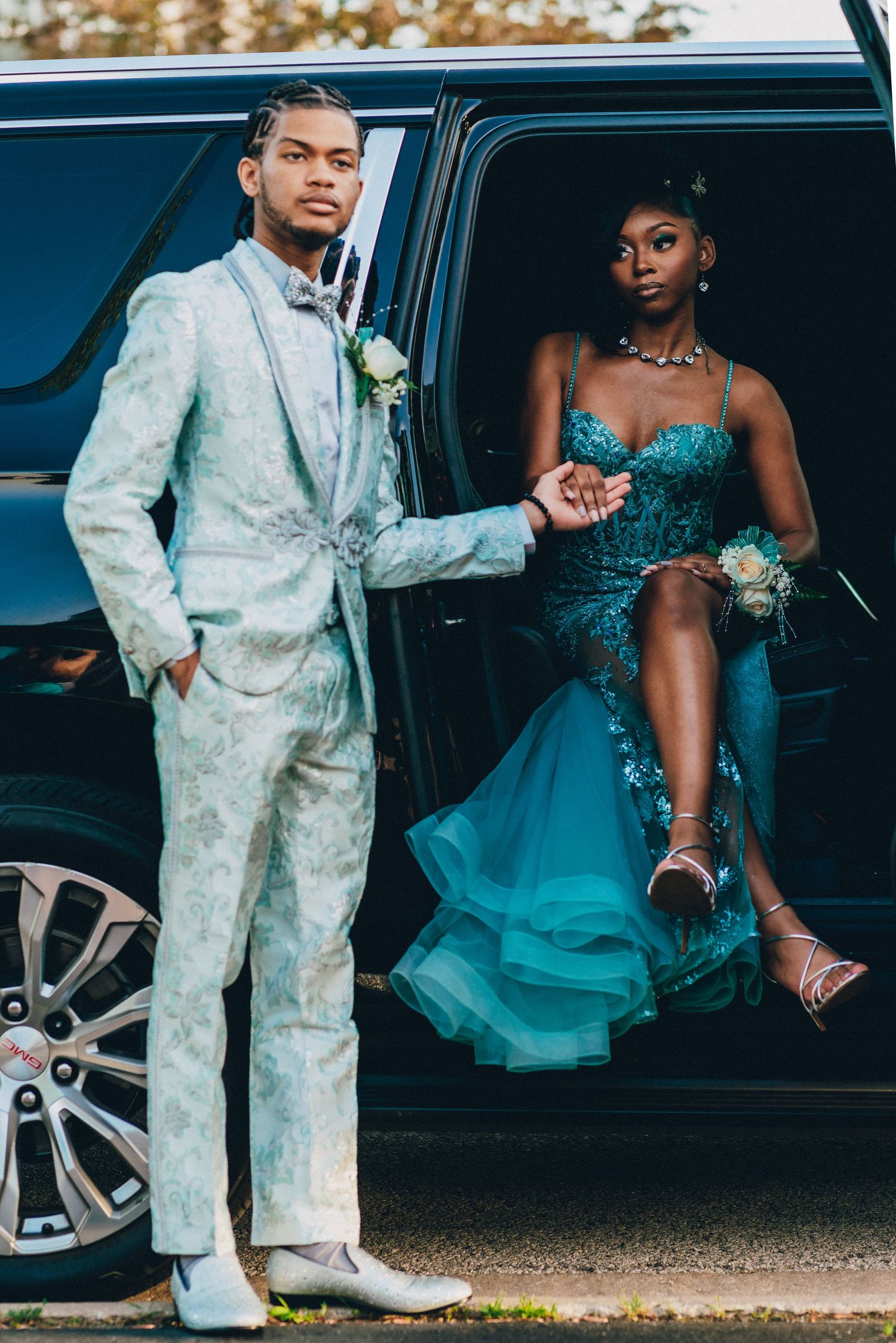 A couple in formal attire poses by a black car, with a white suit and turquoise gown for prom or formal event.