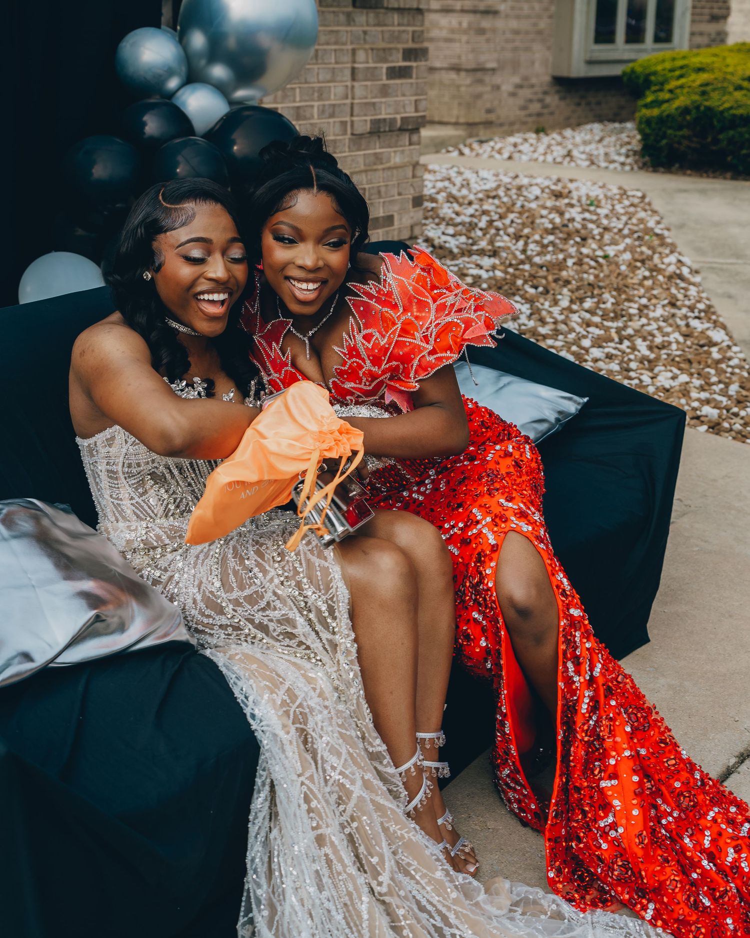 Two friends in elegant dresses share a joyful moment on a couch at a formal event.