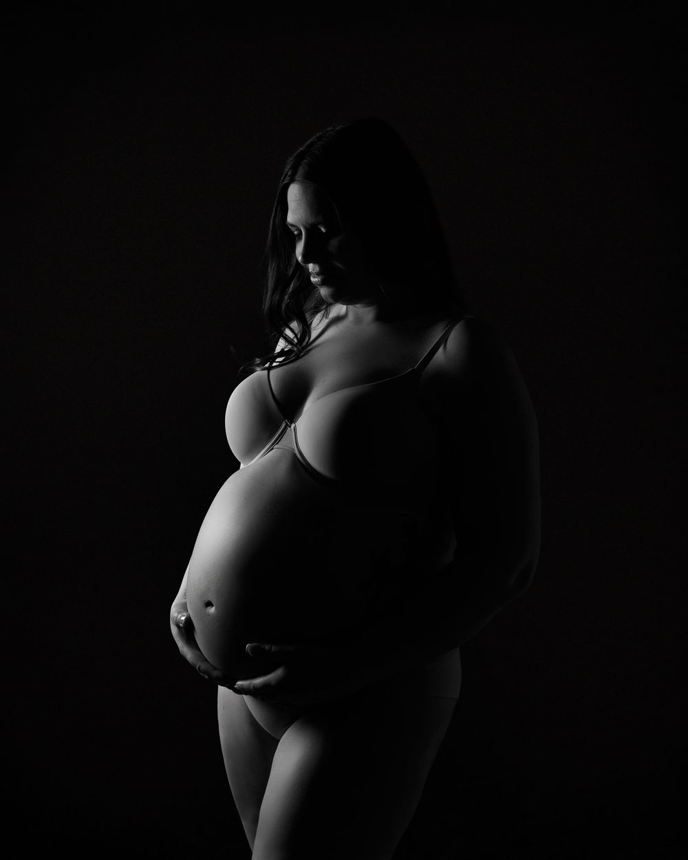 Artistic black and white maternity silhouette photograph showing a pregnant figure against dark background.
