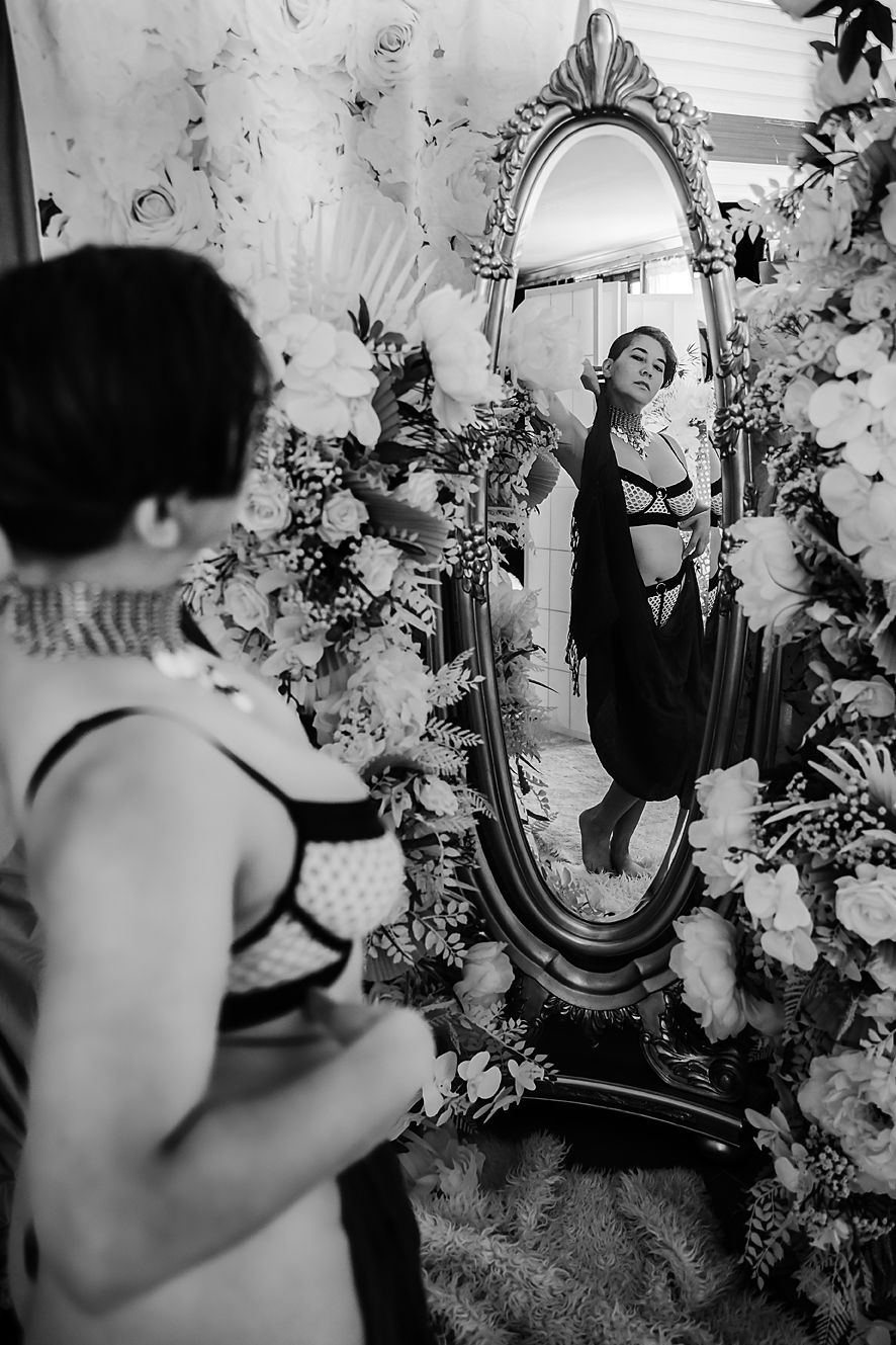 A vintage black and white photograph showing a reflection in an ornate mirror surrounded by floral wallpaper.
