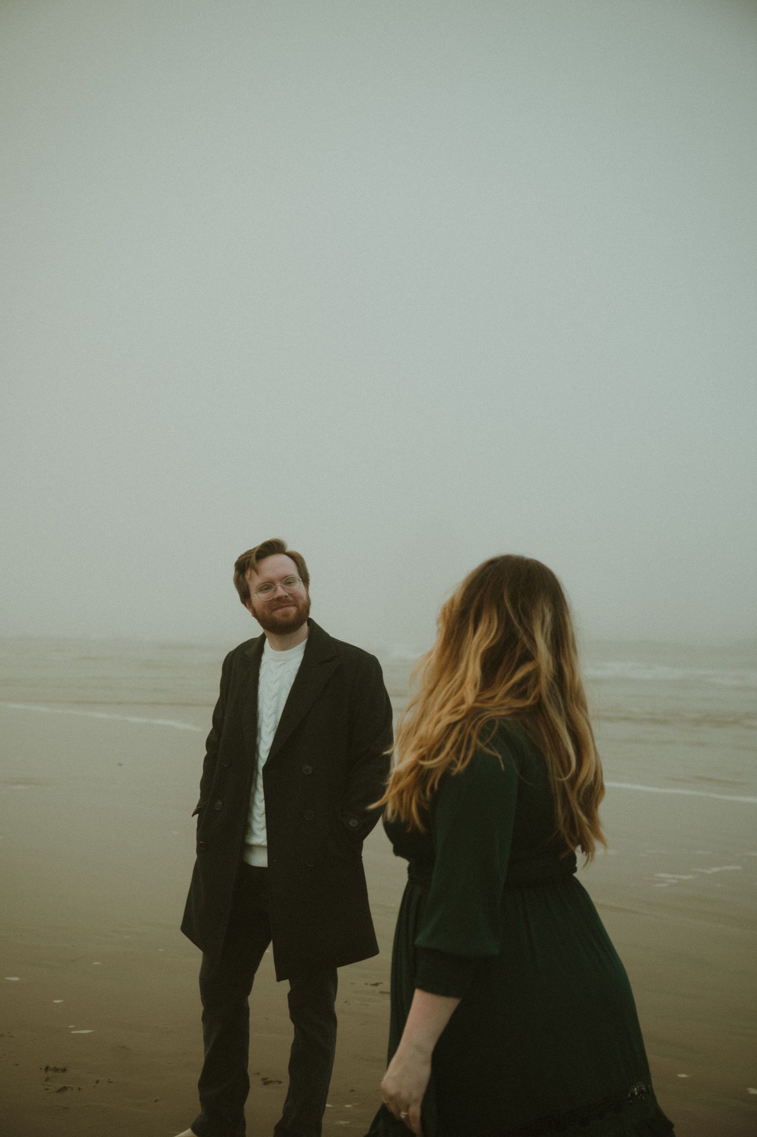 A couple in dark coats stands on a misty beach during an atmospheric photoshoot.