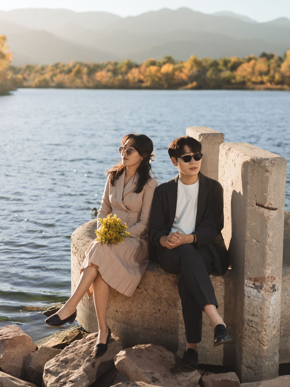 A couple relaxes on concrete blocks by a serene lake during golden hour, creating a romantic coastal scene.