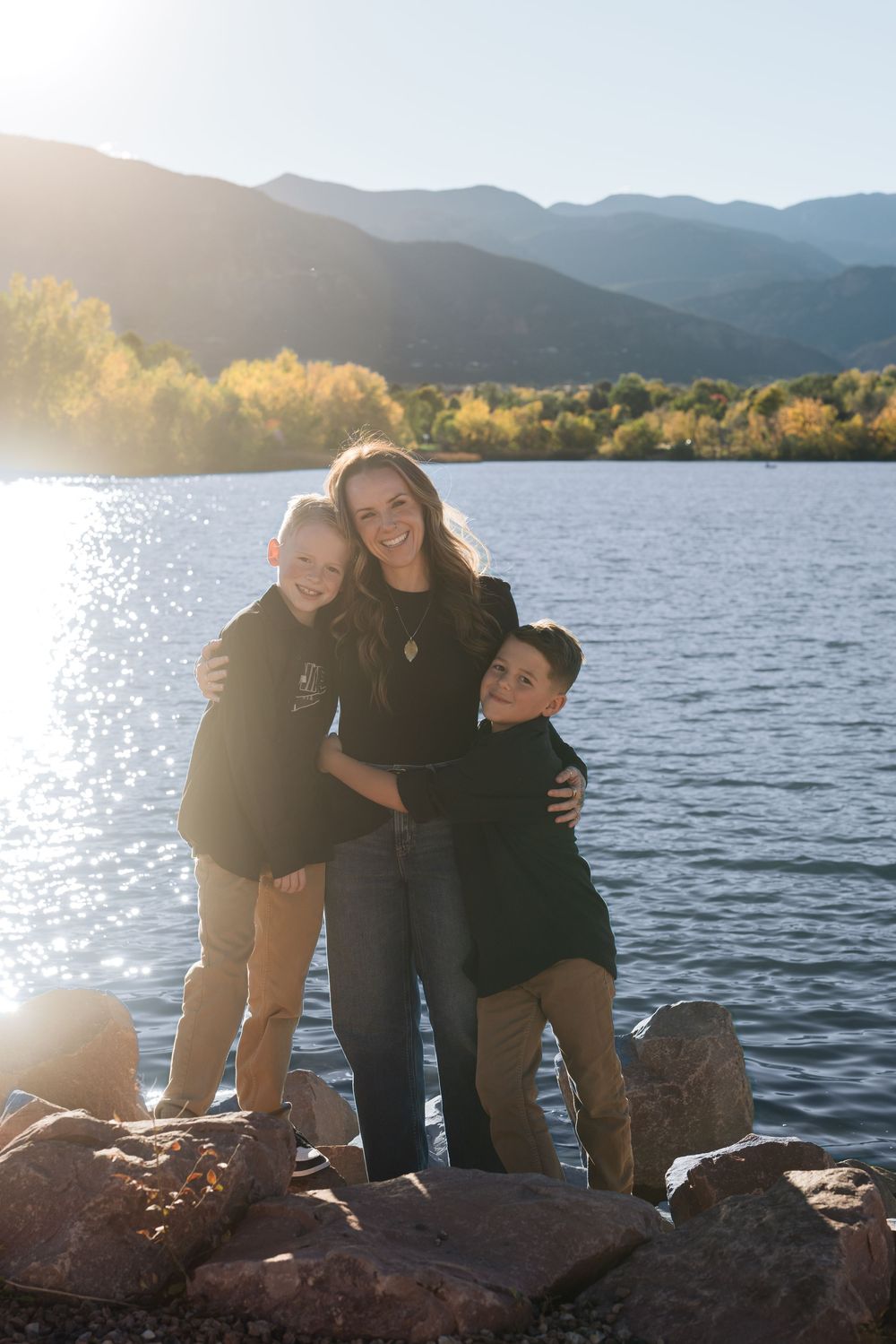 Three people pose together on rocky shoreline of mountain lake at sunset with golden light shining through.