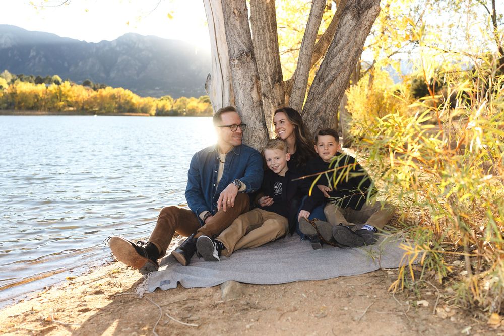 Three people and a dog sit together on a blanket by a lake shore during golden hour with autumn foliage in the background.