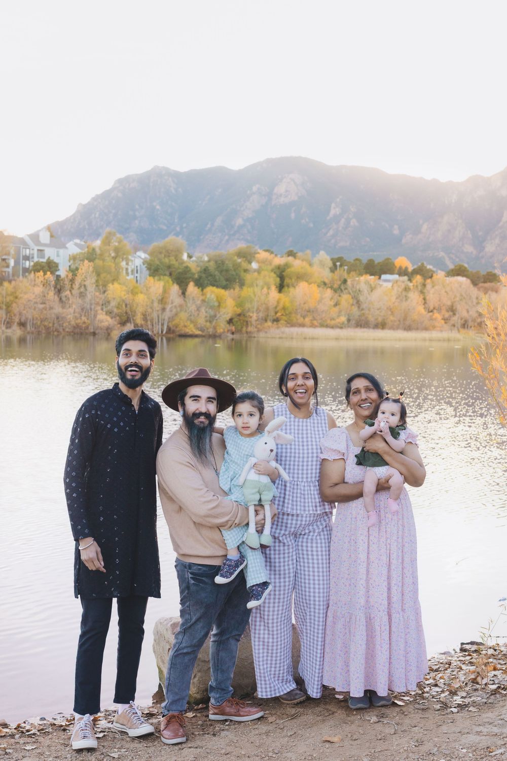 A group of people stand together by a scenic lake with mountains and autumn trees in the background.