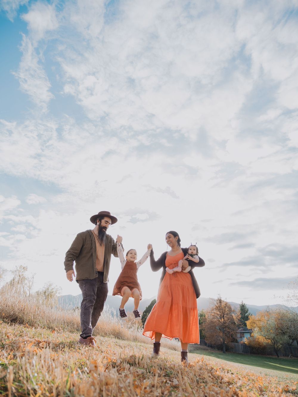 Family holding hands while walking through a golden autumn field against a cloudy sky at sunset.
