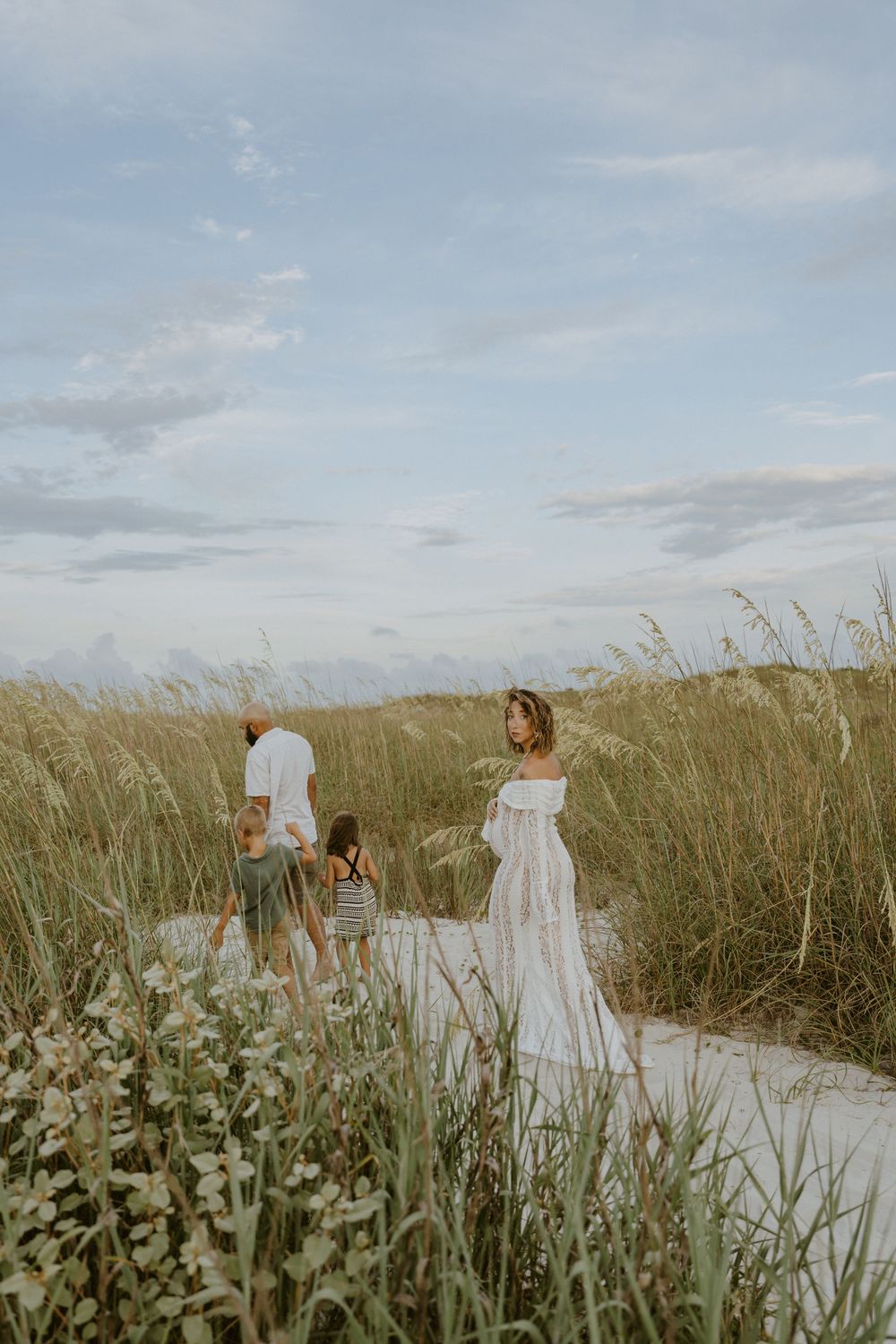 Family walking together through tall beach grass along a sandy coastal path at sunset.