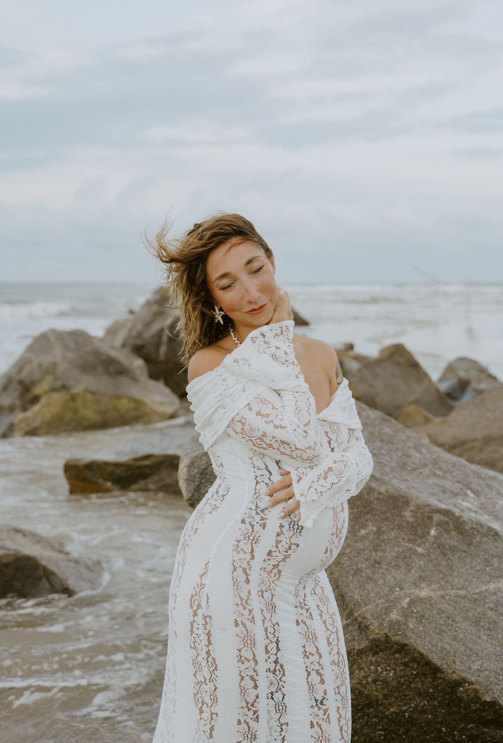 A woman in a white lace dress poses gracefully on rocky coastal rocks at the beach.