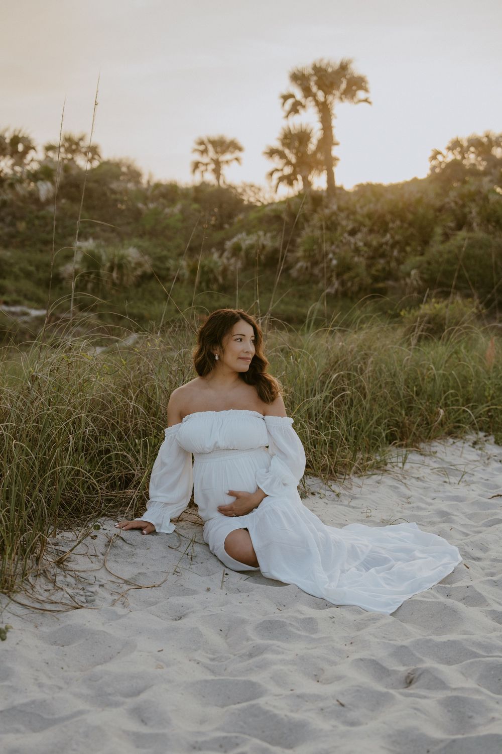 A figure in a flowing white dress reclines on sandy beach dunes at sunset with palm trees in the background.