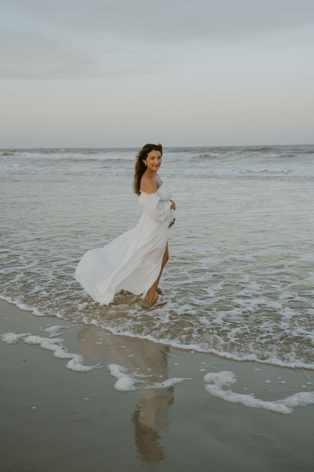 A flowing white dress catches the breeze as waves roll onto a sandy beach at sunset.