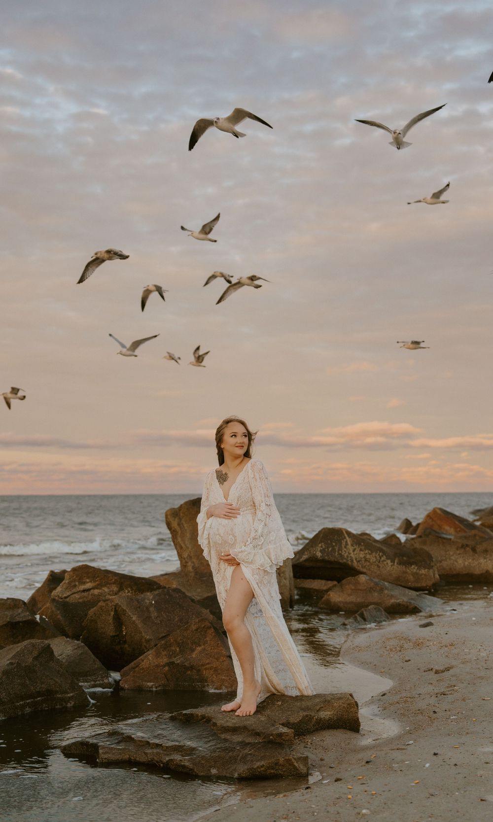 A figure in white lace stands on coastal rocks at sunset while seagulls soar overhead against a dusky sky.