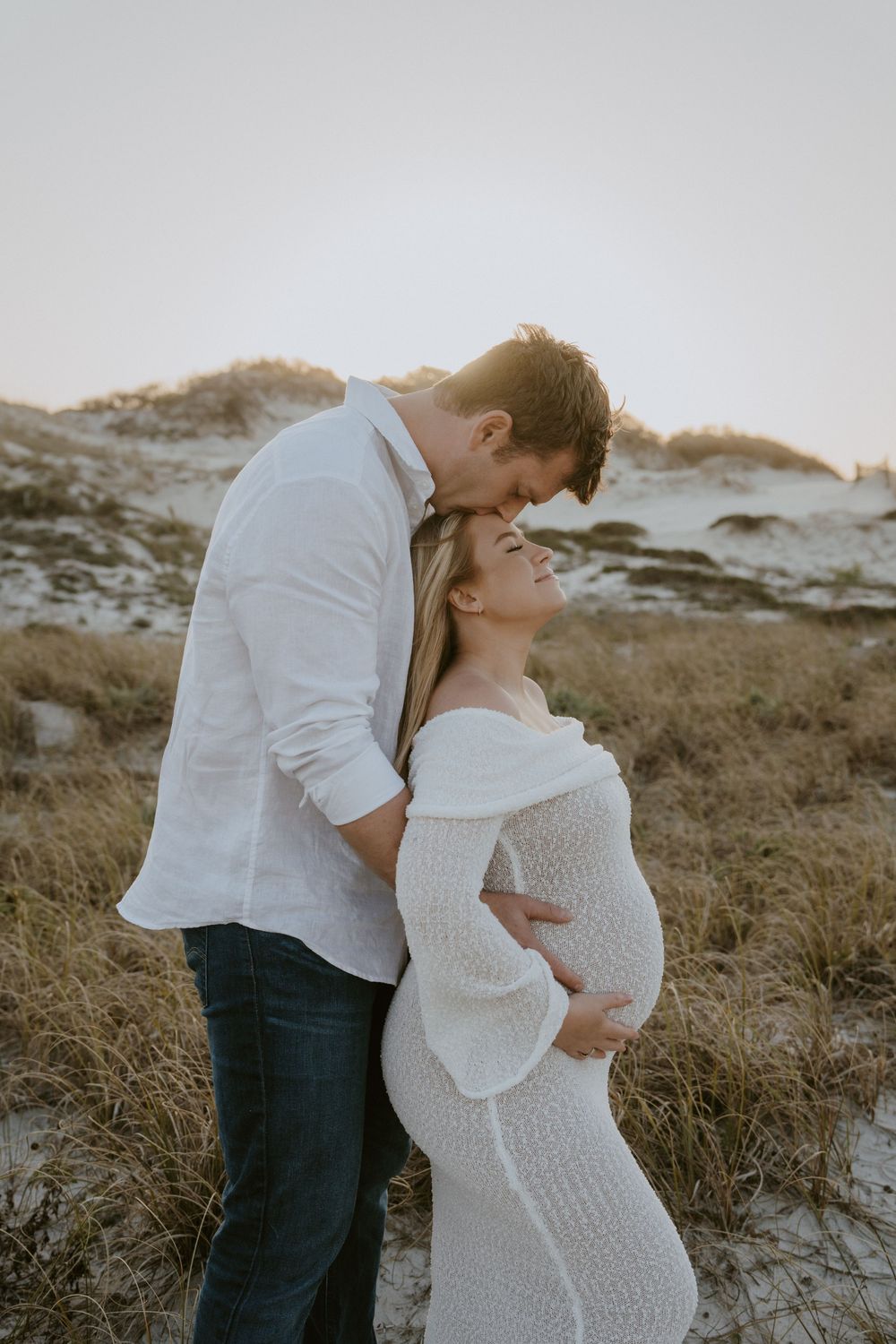 Romantic beach photoshoot featuring a couple in white embracing against a sunset backdrop.