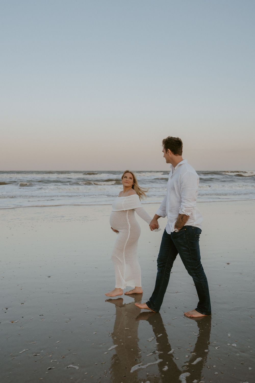A couple walks barefoot along a misty beach at sunset with waves rolling in the background.