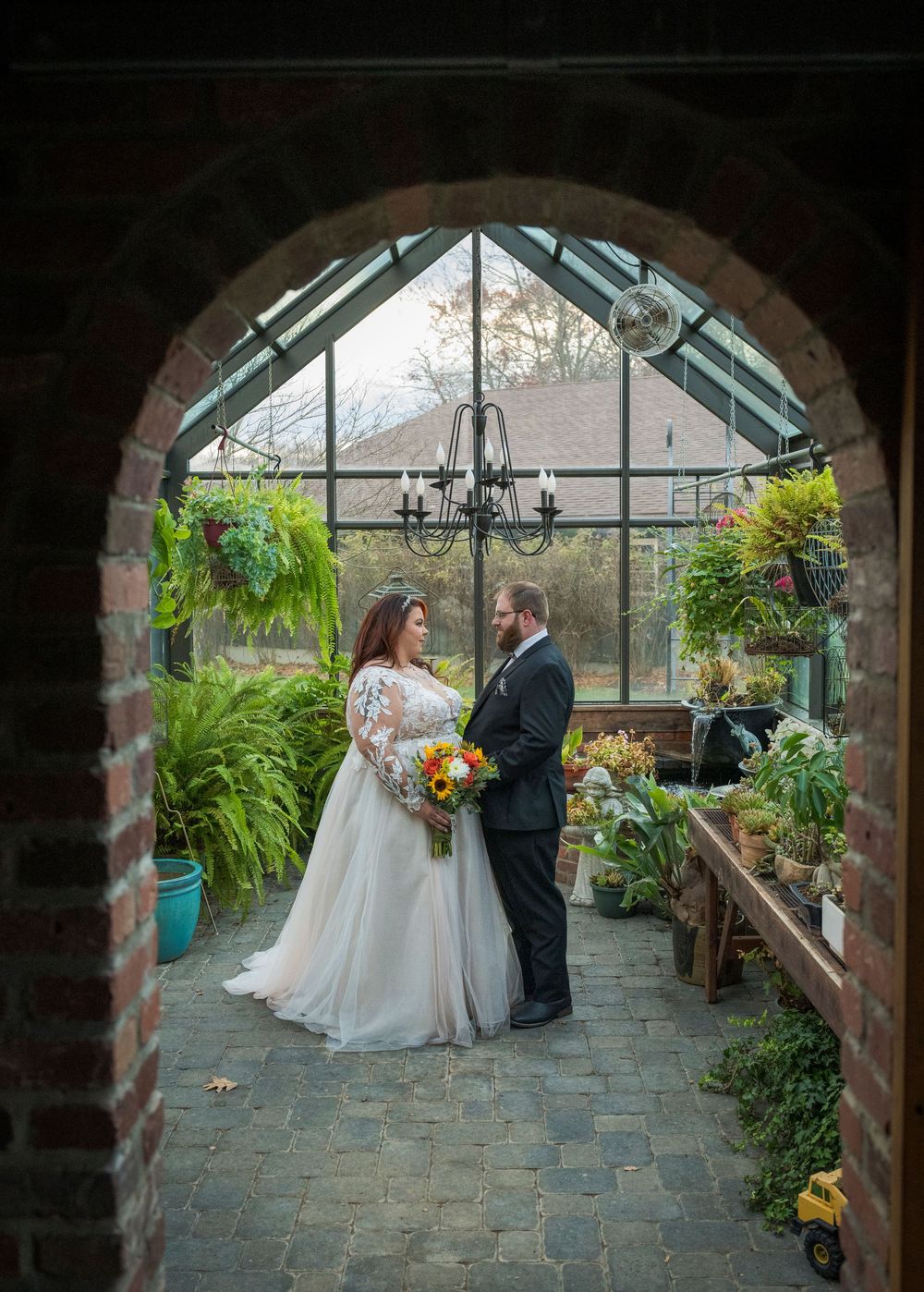 A couple shares an intimate moment in a greenhouse conservatory framed by a stone archway and surrounded by lush plants.
