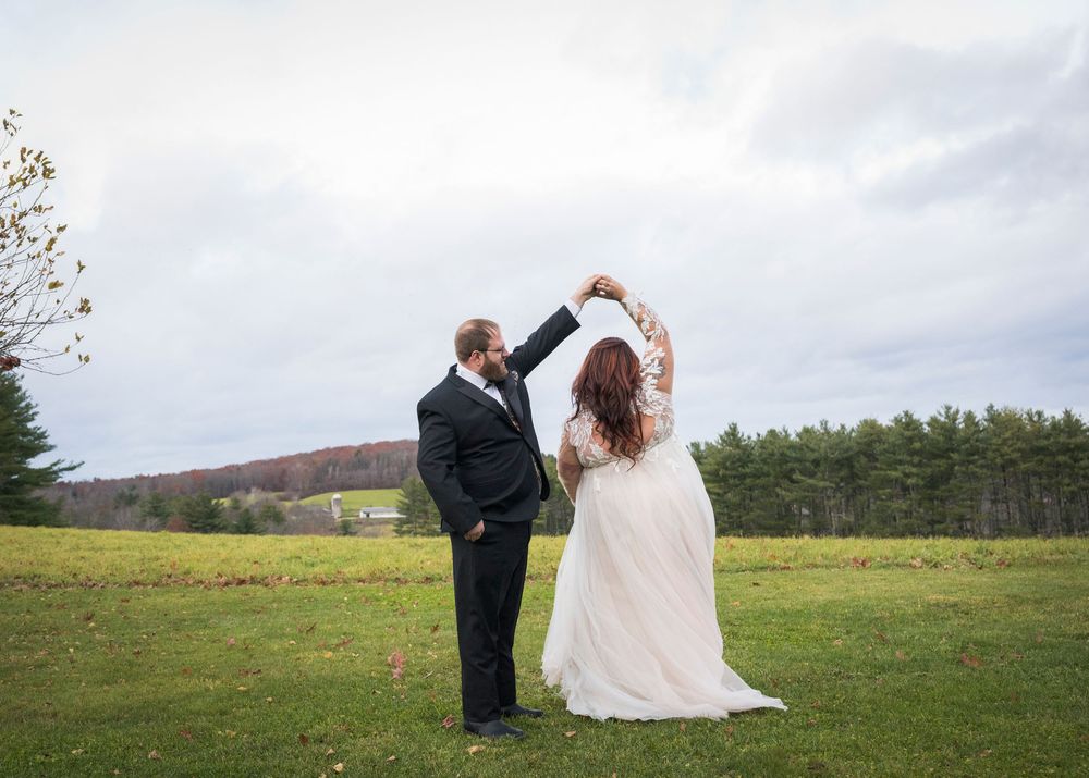 Couple in wedding attire throws confetti in a scenic meadow with mountains and cloudy skies in the background.