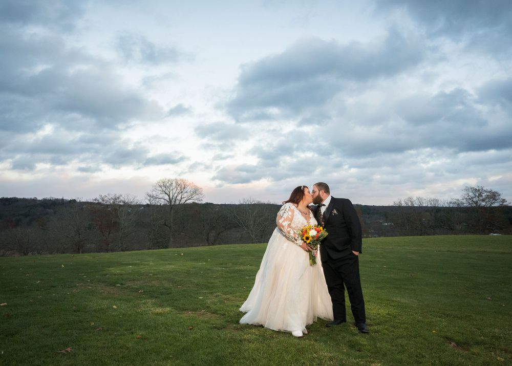 A couple in wedding attire shares a romantic moment in a scenic green field under dramatic cloudy skies.
