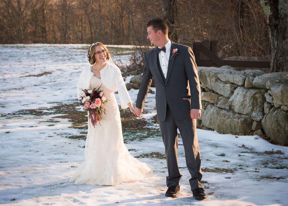 A couple walks together in the snow holding hands with a bridal bouquet against a winter landscape backdrop.