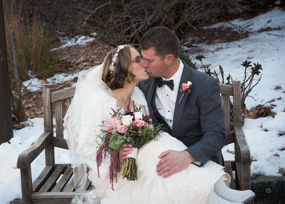 A couple shares a romantic winter moment on a rustic wooden bench surrounded by snow.