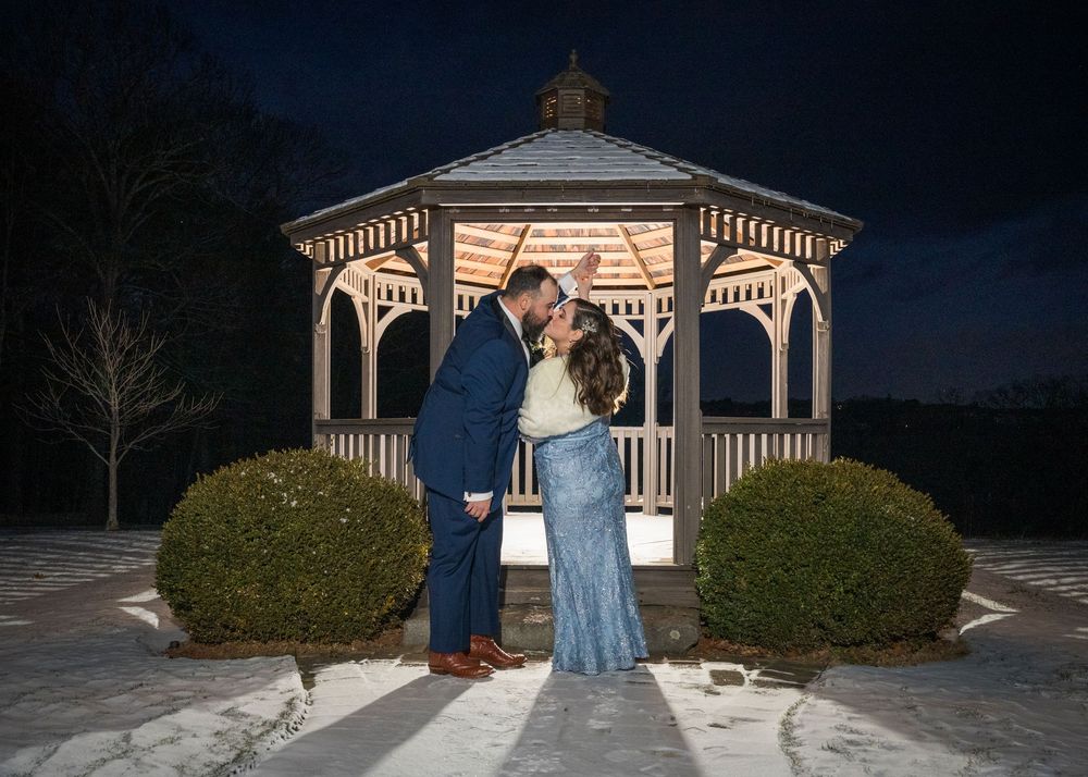 A romantic nighttime scene at an illuminated white gazebo with two figures embracing between manicured shrubs.