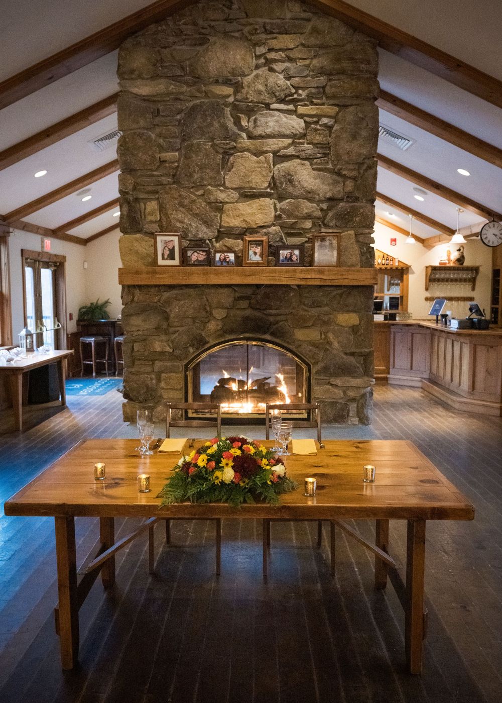 Rustic wooden dining table with floral centerpieces sits beneath stone fireplace and exposed wooden beams in elegant venue.