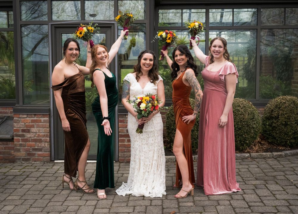 Wedding party poses together in formal attire with sunflower bouquets outside glass building entrance.