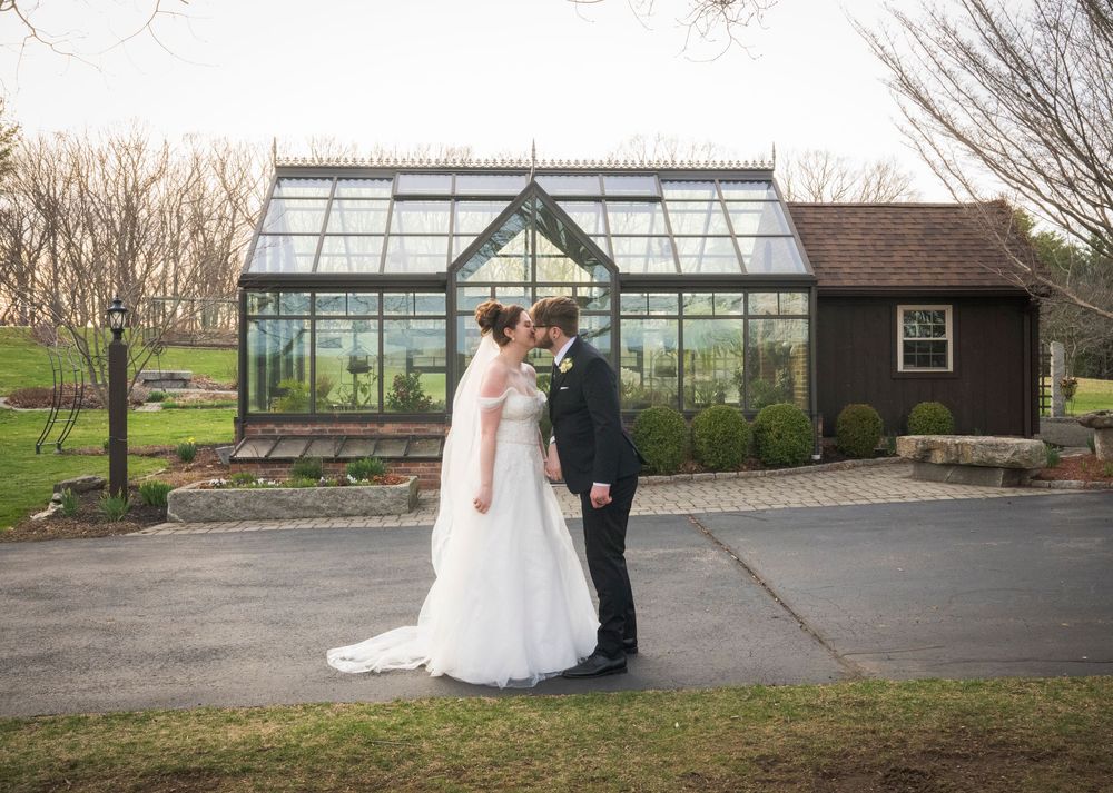 A couple shares a romantic moment in front of a glass greenhouse with a rustic wooden shed in the background.