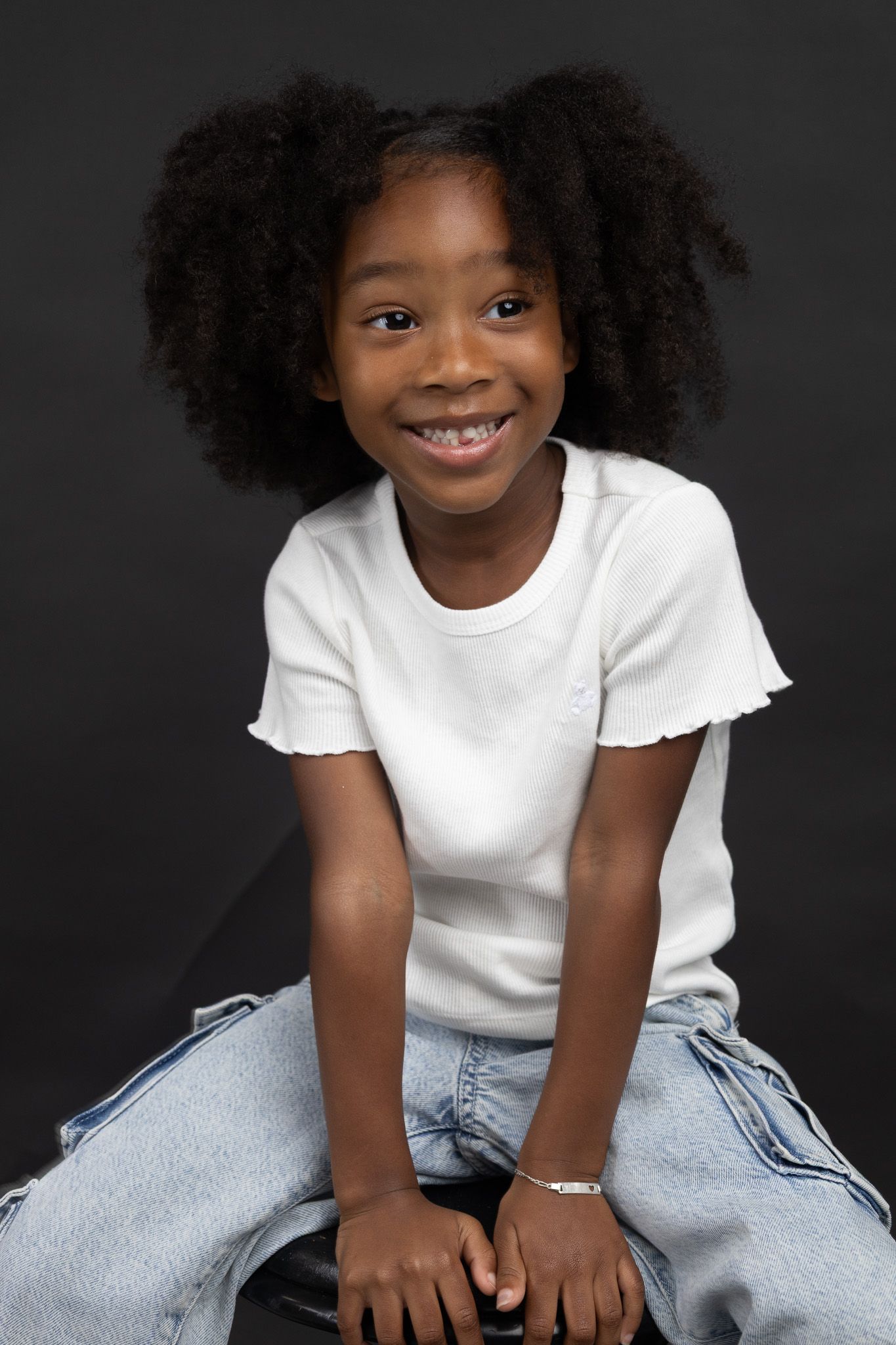 A child in a white t-shirt and jeans poses against a dark background with natural curly hair and a bright smile.