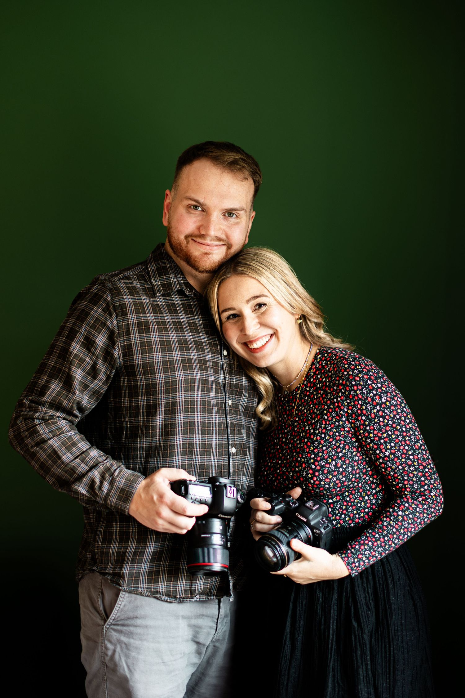 A smiling male and female photography duo pose together holding DSLR cameras against a dark green background.