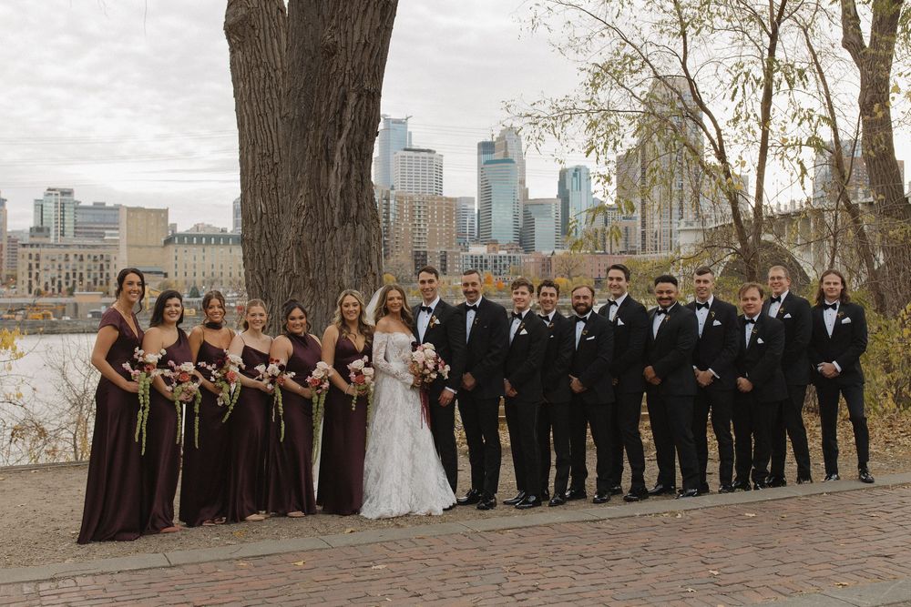 Wedding party in formal attire poses outdoors with city skyline backdrop on autumn day.