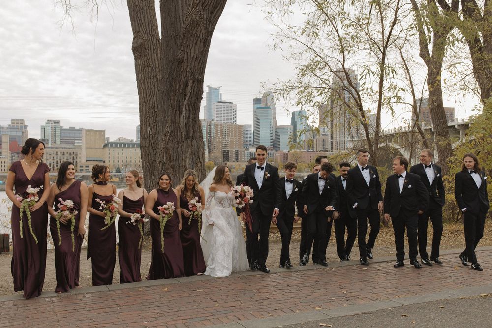 Wedding party in formal attire walking along a city path with autumn trees and urban skyline in the background.
