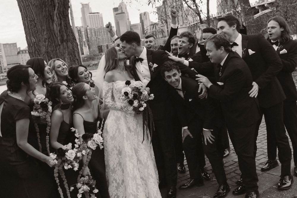 Wedding group photo with burgundy and black attire against city landscape.