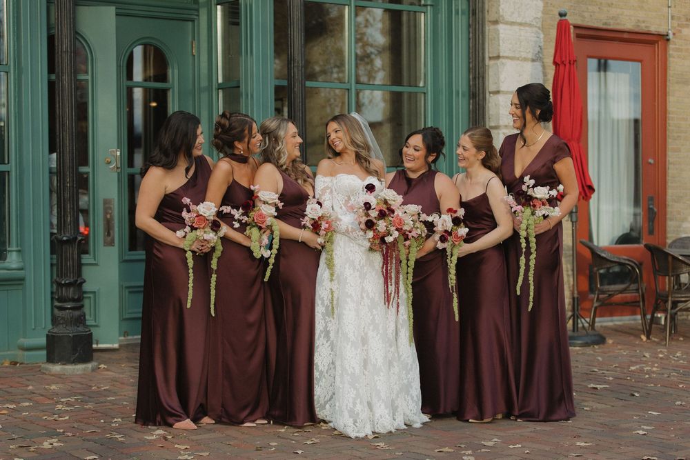 A wedding party in brown and white dresses poses along a historic brick street with colorful buildings in the background.