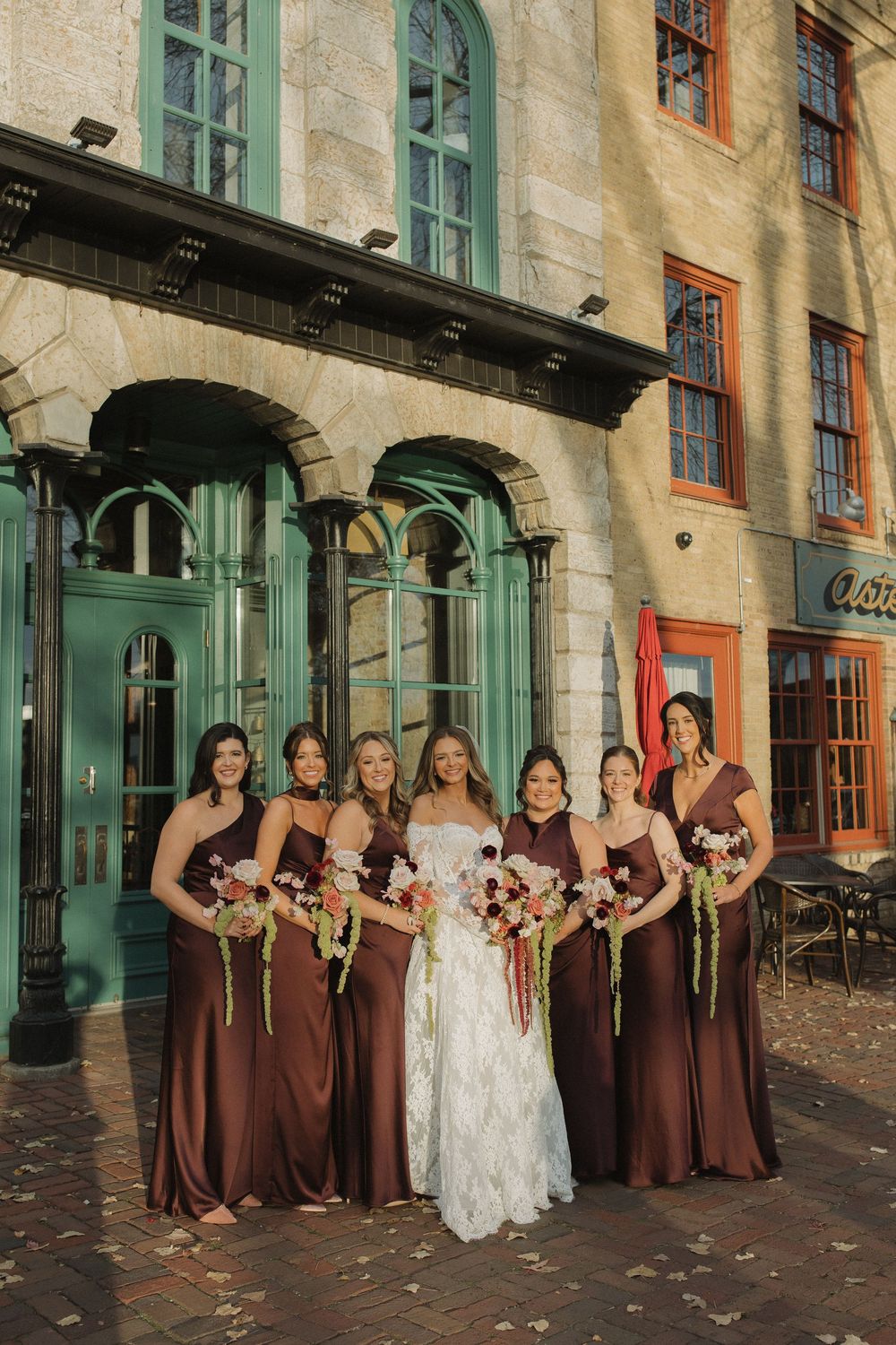 Wedding party in chocolate brown dresses poses in front of historic building with turquoise shutters.