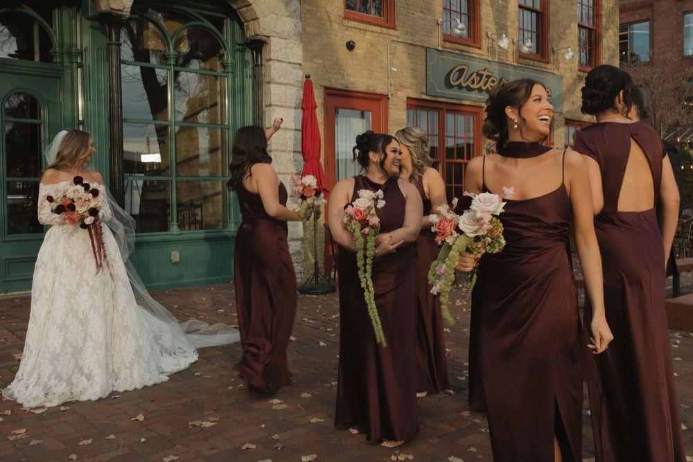 Bridal party walks along brick building exterior in brown dresses holding pink bouquets.