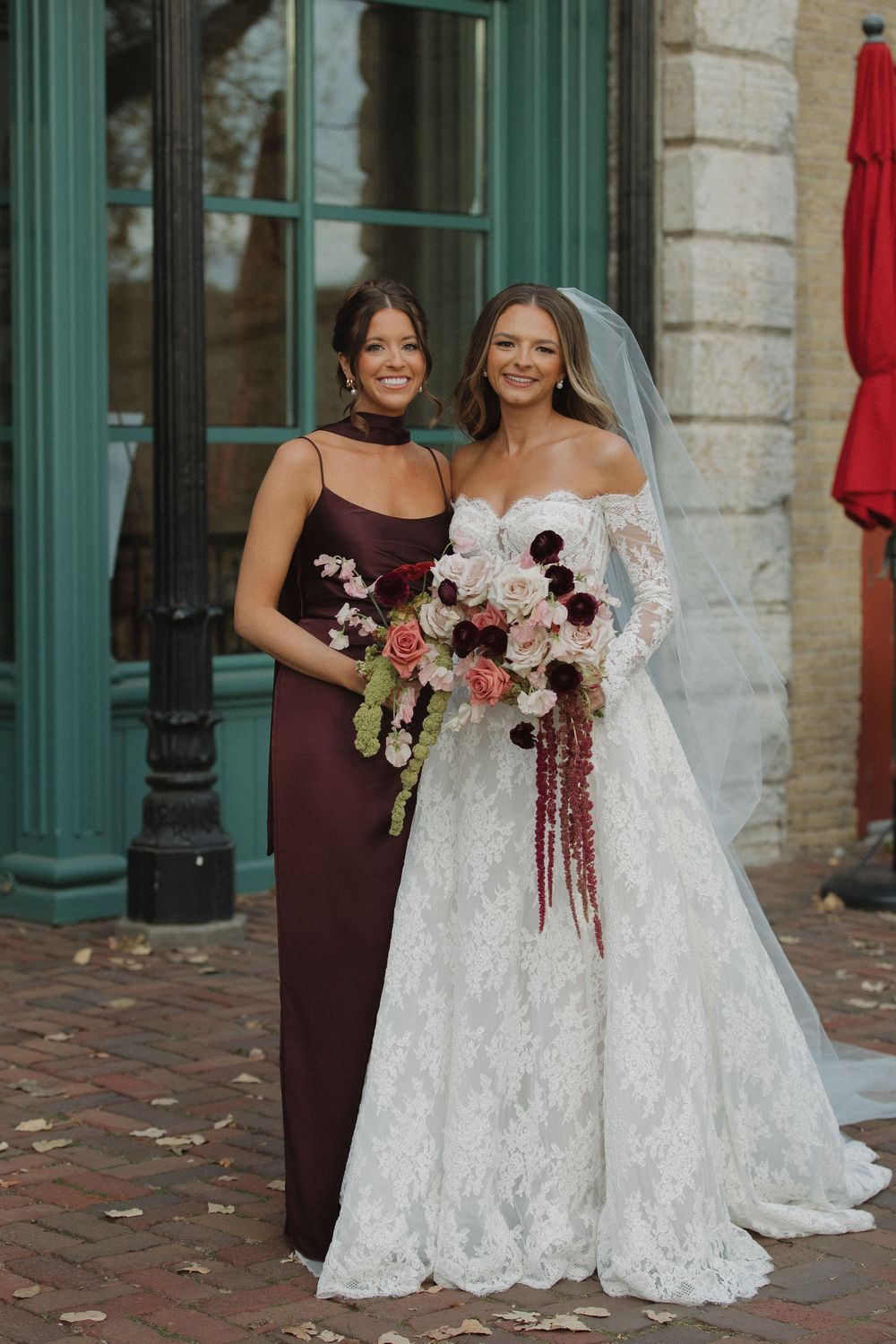 A bride in a lace off-shoulder gown and bridesmaid in burgundy dress pose with blush and burgundy floral bouquets.