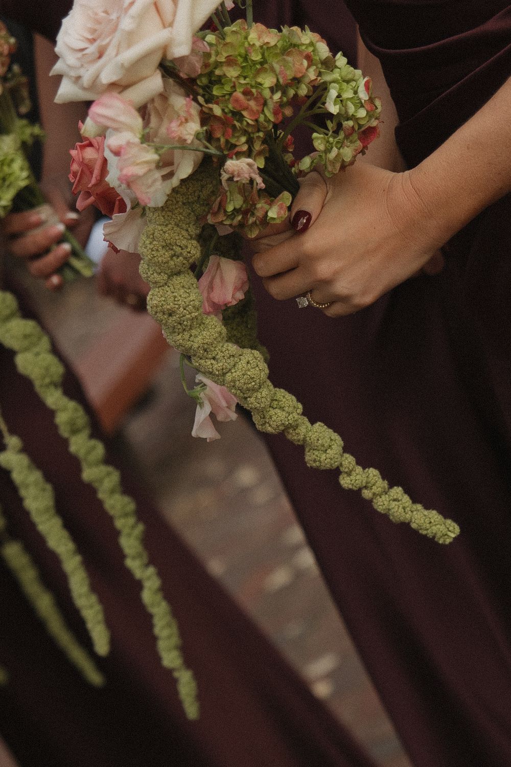 Close-up of pink and green floral wedding bouquets with cascading amaranthus against brown dresses.