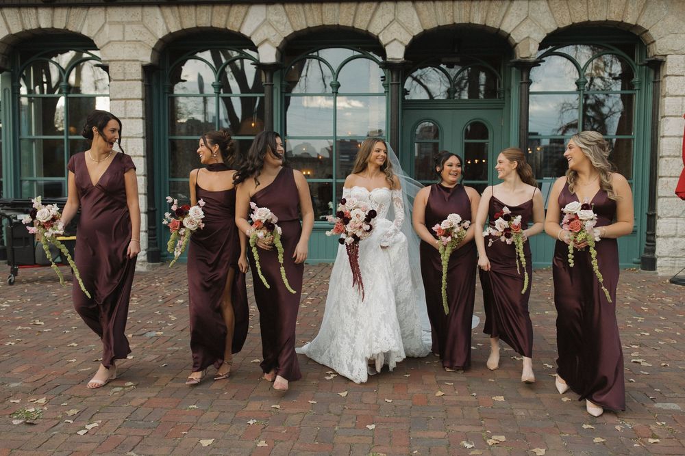 Wedding party in burgundy dresses and suits poses together on brick patio with scattered flower petals.