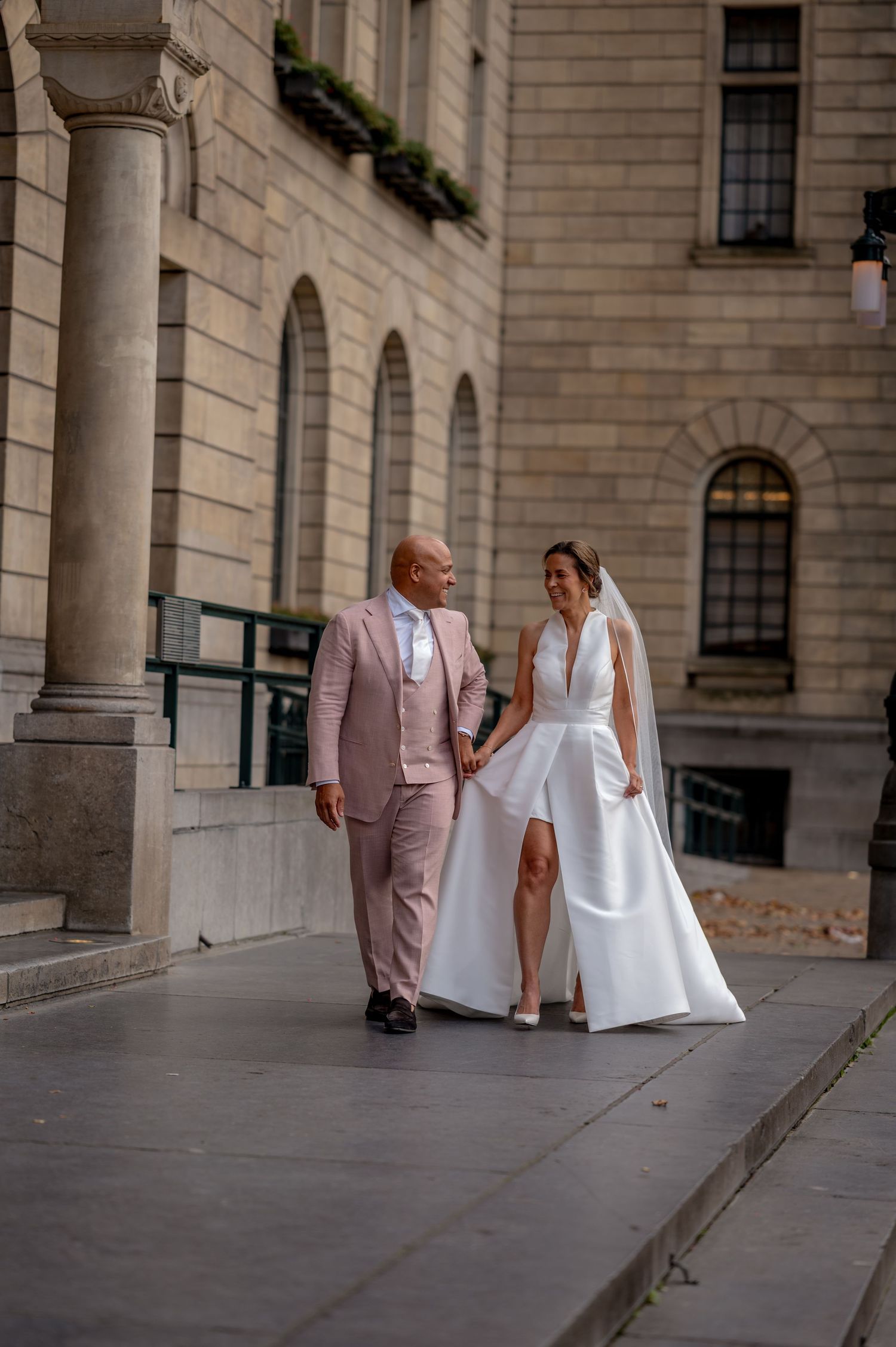 A couple in wedding attire walks down a stone-paved walkway in Rotterdam outside a historic building.