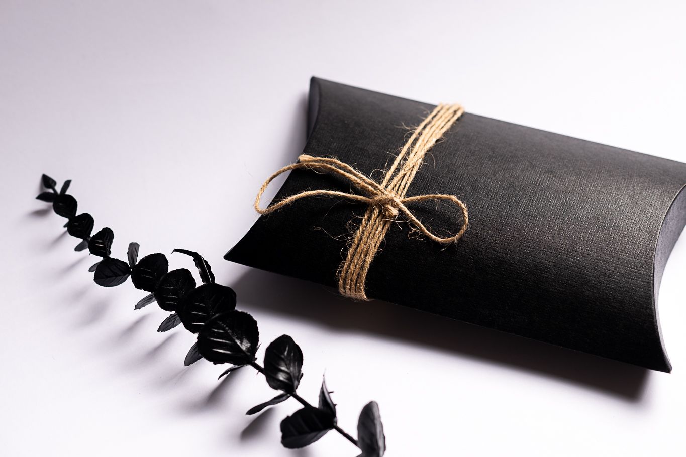 Black pillow box gift wrapped with twine bow and eucalyptus sprig on gray background.