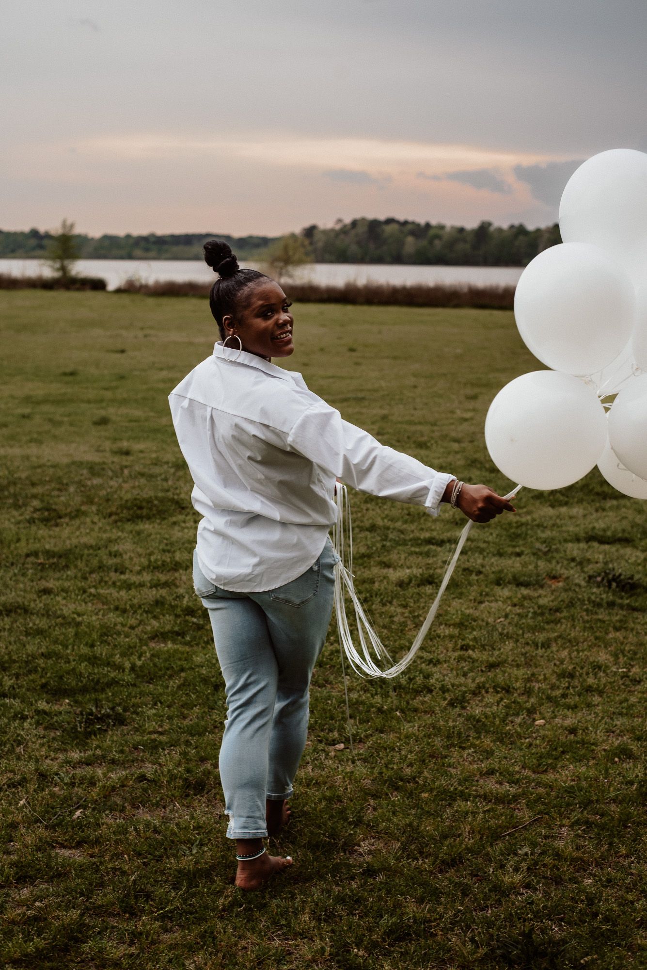 Person in white shirt and jeans dancing with white balloons in a grassy field at dusk.