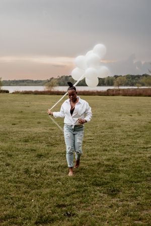 Person runs playfully through an open field while holding white balloons.